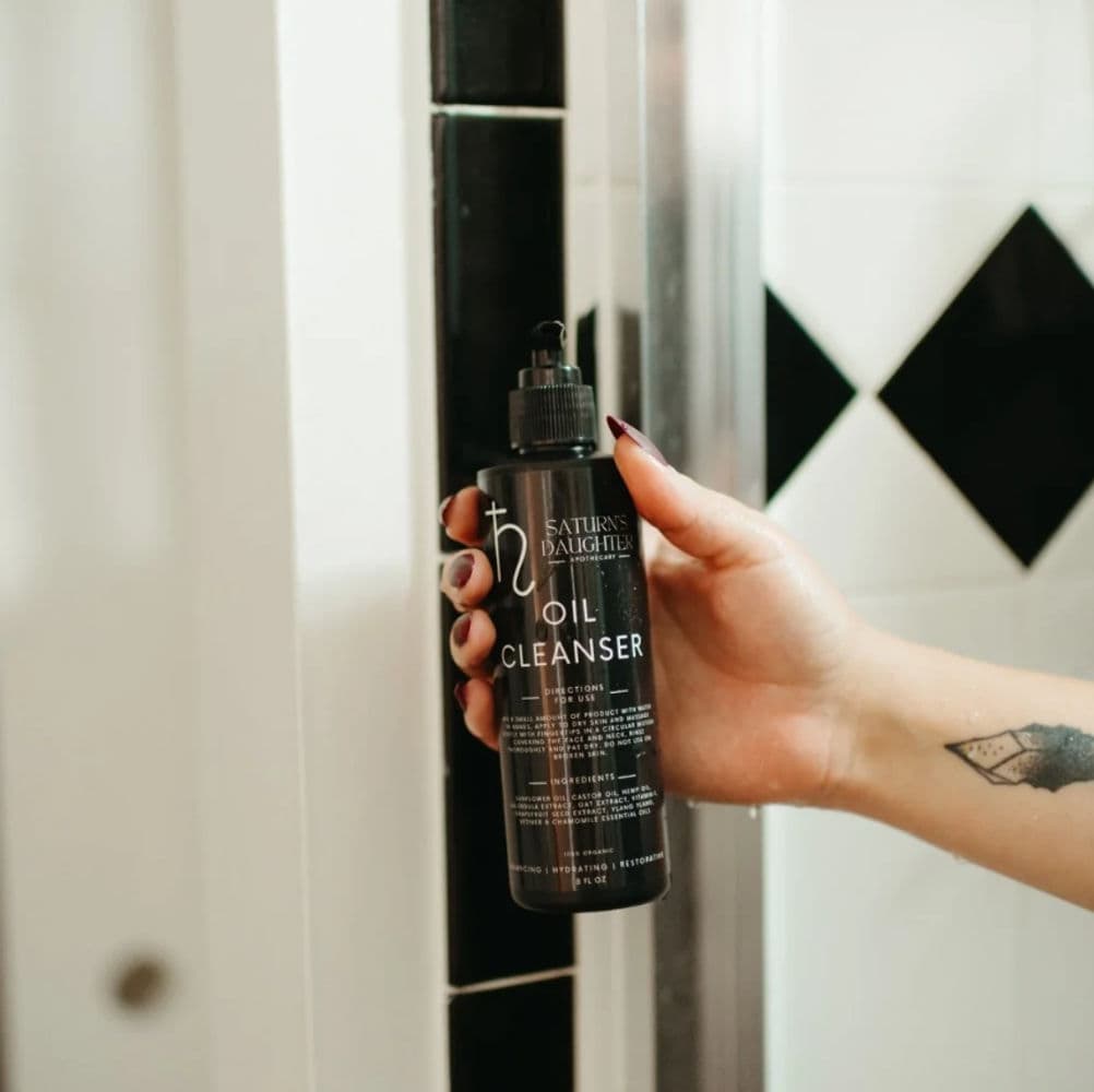Product photography of a cosmetic oil cleanser bottle held in hand against a minimalist interior backdrop.