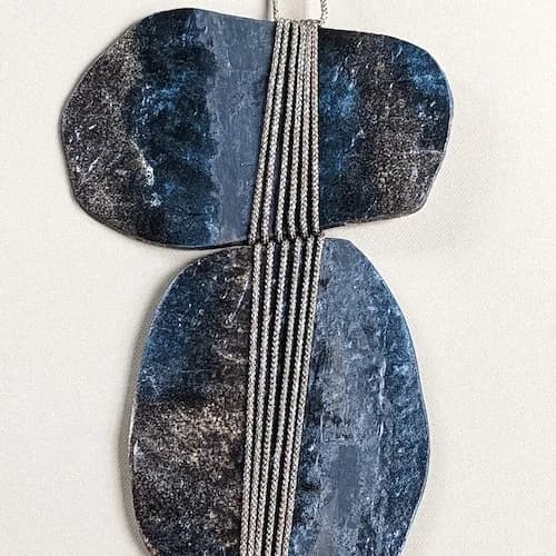Abstract stone sculpture with geometric form and linear textile accent suspended vertically.