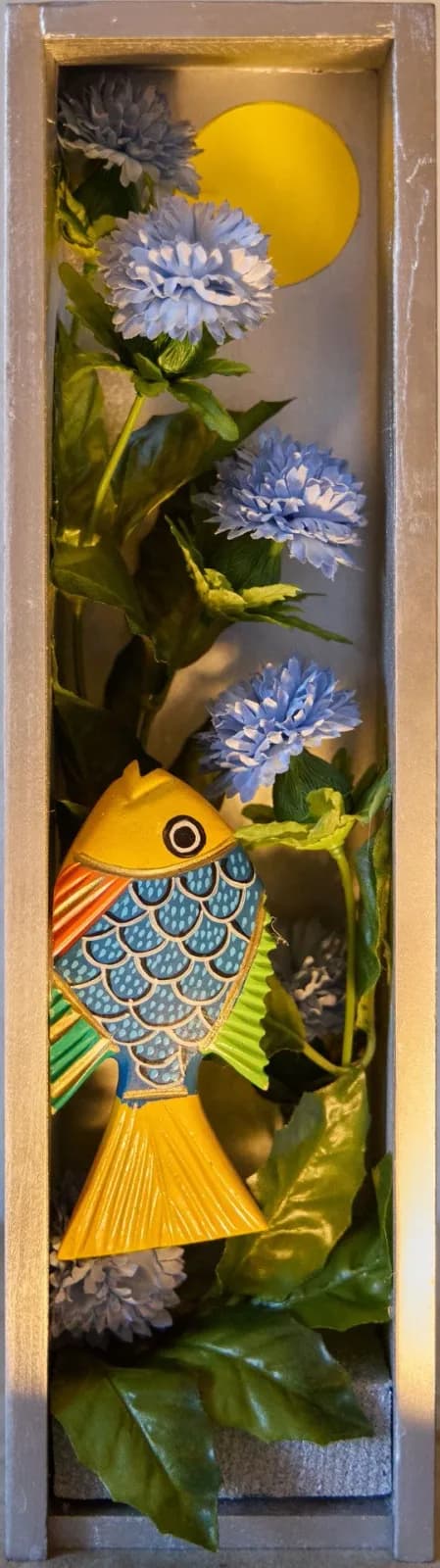 Hand-carved wooden fish and blue flowers arranged in a framed shadow box composition.