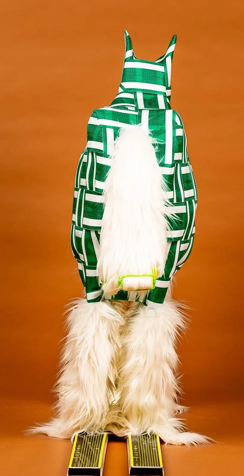 A llama figure wrapped in green and white geometric textile strips stands against a burnt-orange background.