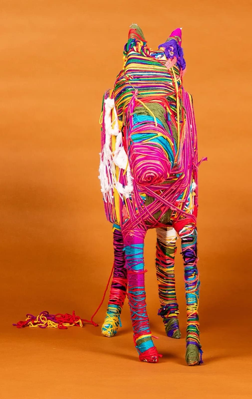 Vibrant yarn-wrapped sculptural form in rainbow colors stands against a warm orange backdrop.
