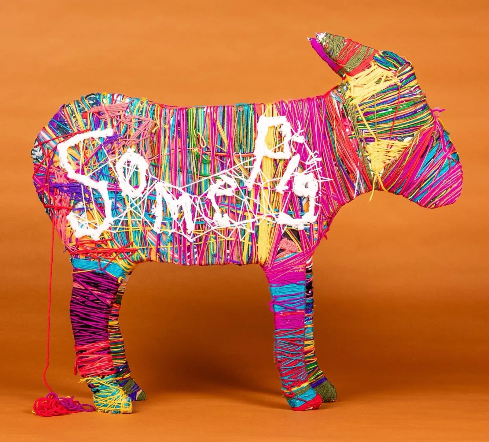 A colorful three-dimensional cow form constructed from vibrant multicolored yarn and fiber against an orange background.