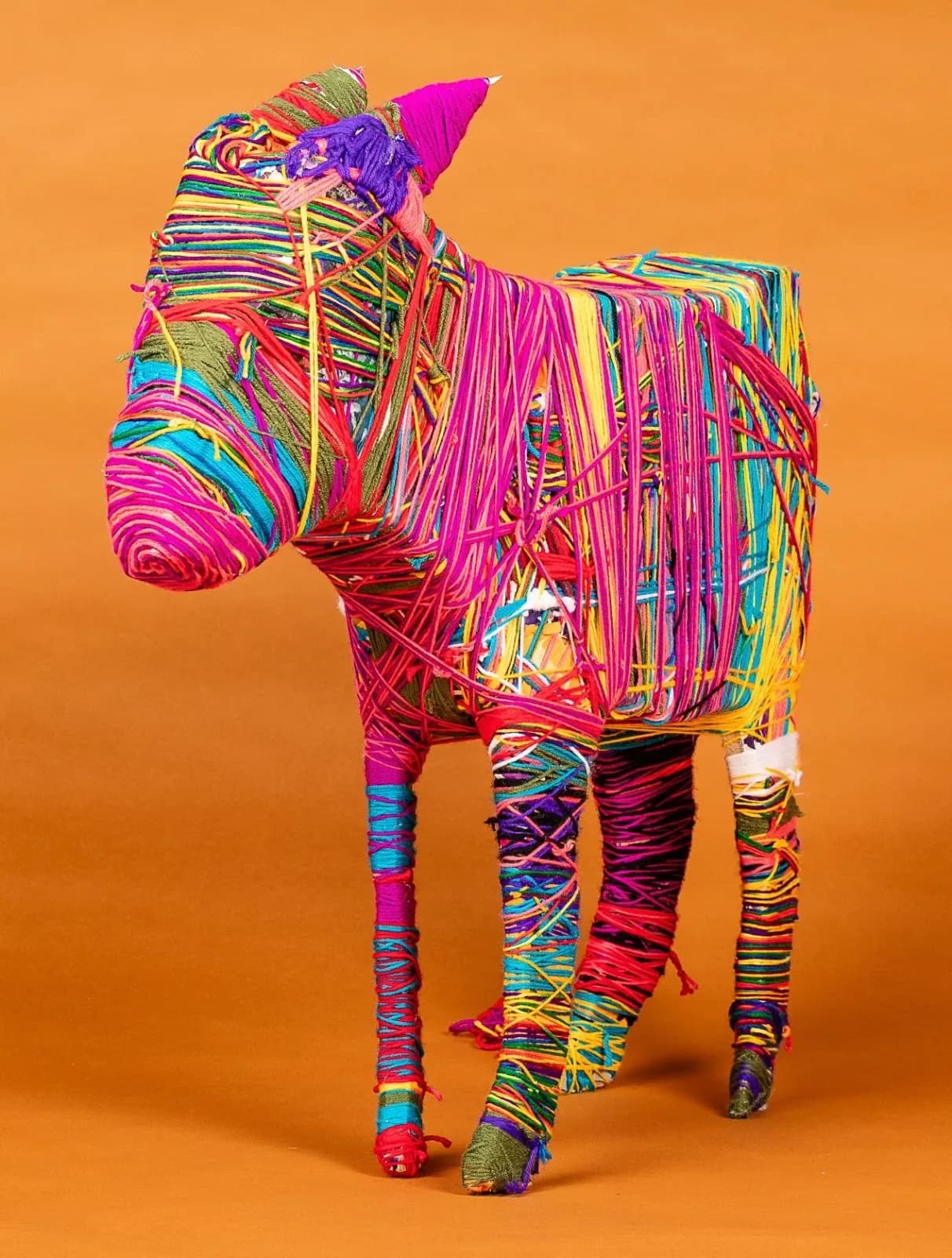 Colorful yarn-wrapped sculptural form in neon pinks, blues, and yellows against an orange background.