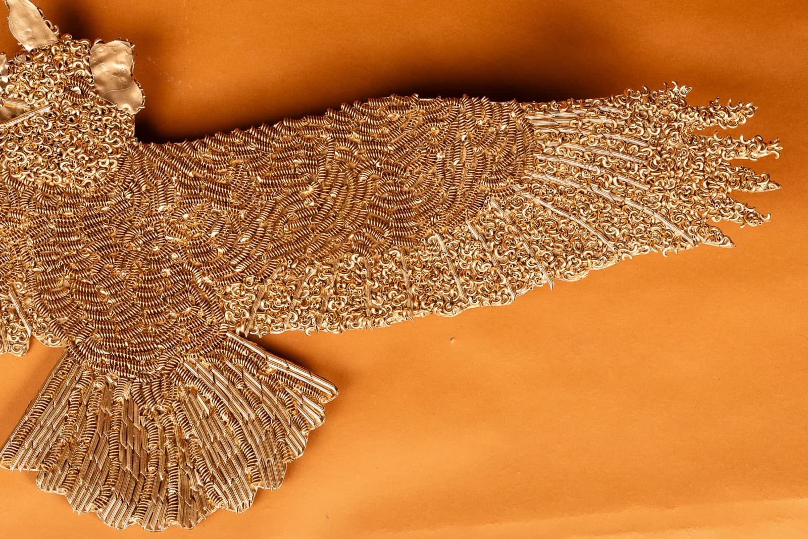 Abstract metal sculpture with intricate textured forms suggesting bird or wing imagery against warm orange background.