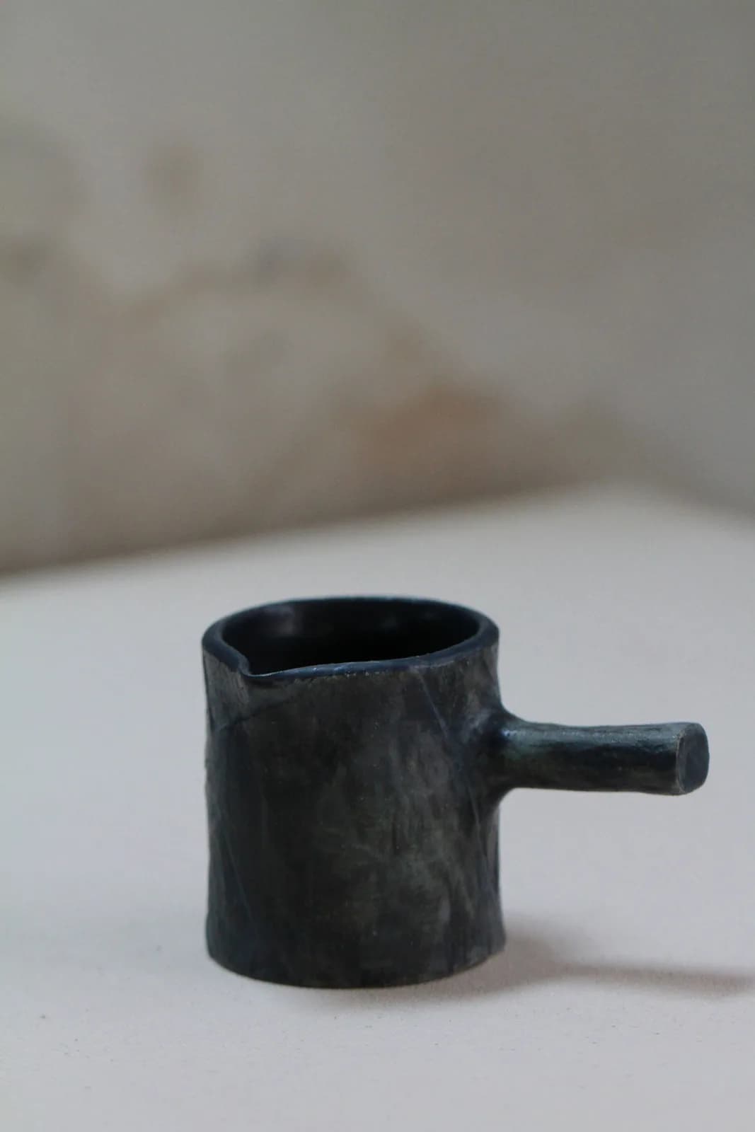Dark glazed ceramic vessel with cylindrical body and side spout on white surface.