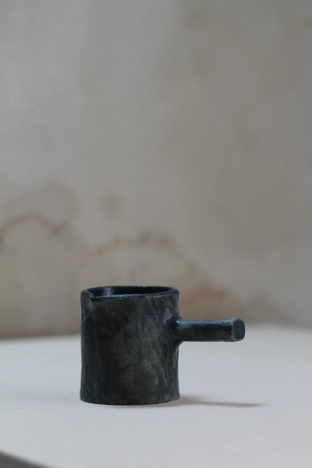 Minimalist dark ceramic vessel with cylindrical body and small spout on neutral background.