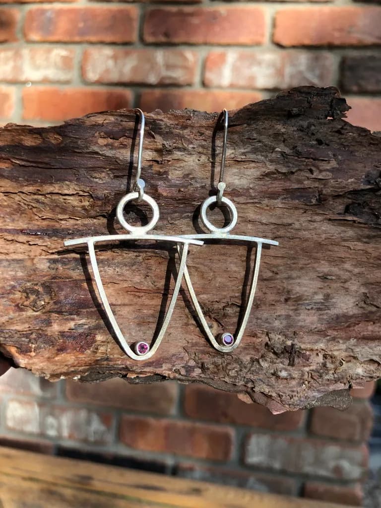 Sterling silver wire earrings with geometric forms and small stone accents, displayed on weathered bark.
