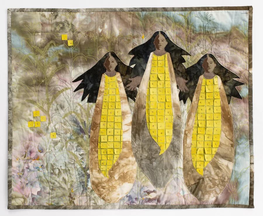 Three winged figures hold oversized yellow corn cobs in a surreal botanical textile work with layered, painterly backgrounds.