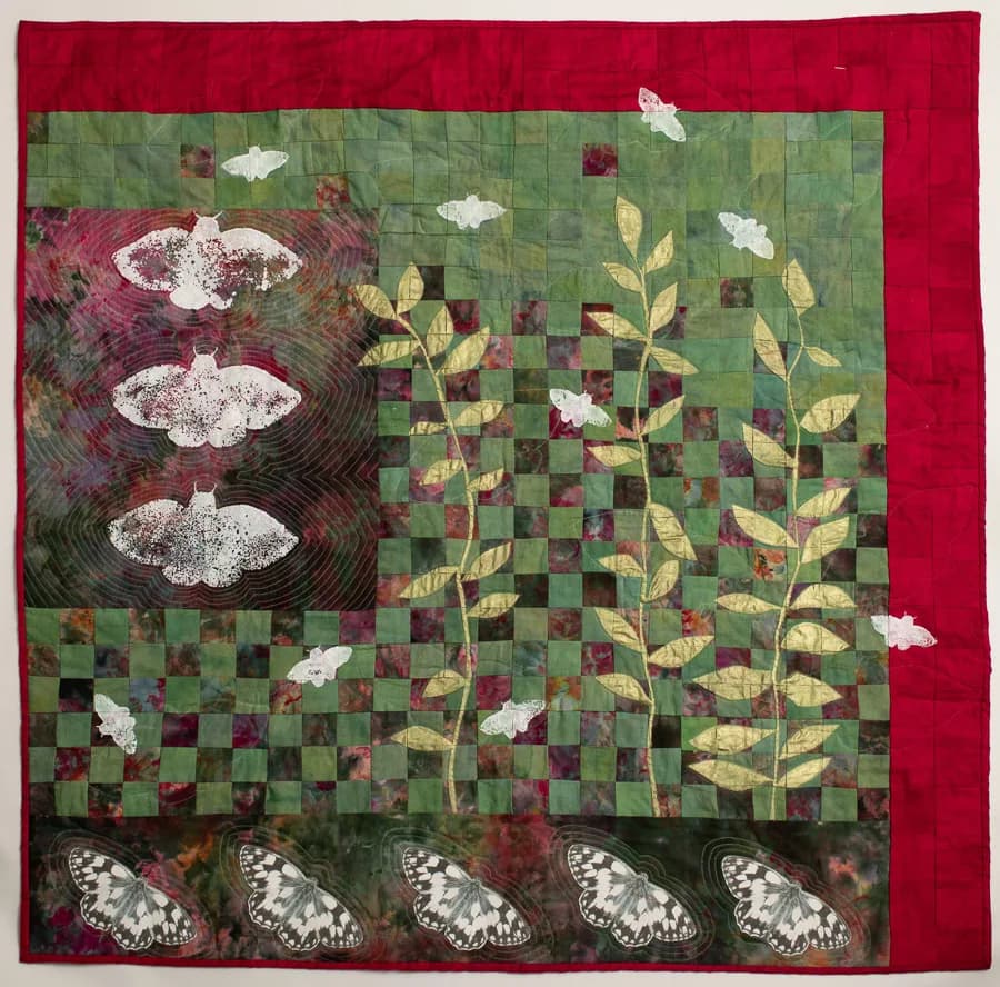 Quilted textile artwork featuring butterflies, moths, flowering plants, and cloud motifs on a checkered green field with red border.
