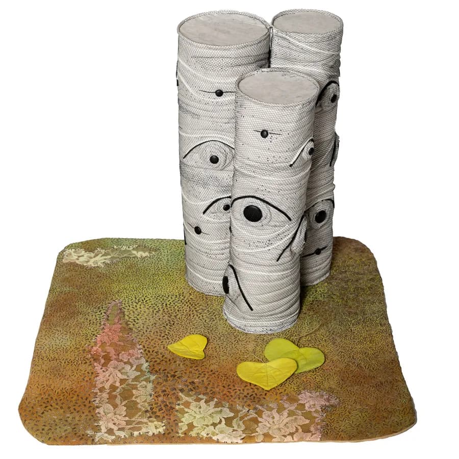 Fiber and textile sculpture featuring wrapped cylinders with drawn eyes and yellow feet on a moss-textured base.