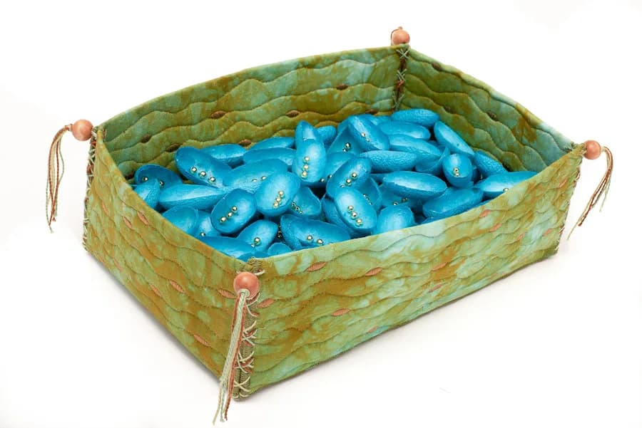 A textile fiber basket with stitched seams holds abstract blue sculptural forms inside.