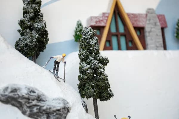 A figure on skis descends a snowy diorama landscape with miniature trees and an alpine cottage in the distance.