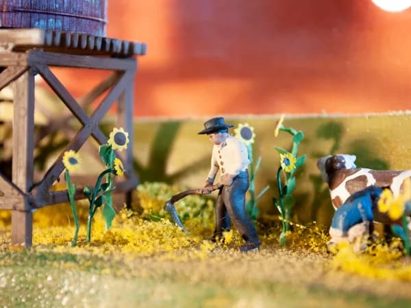 Miniature Western homestead scene with painted metal figures, sunflowers, and wooden structures against burnt sienna sky.