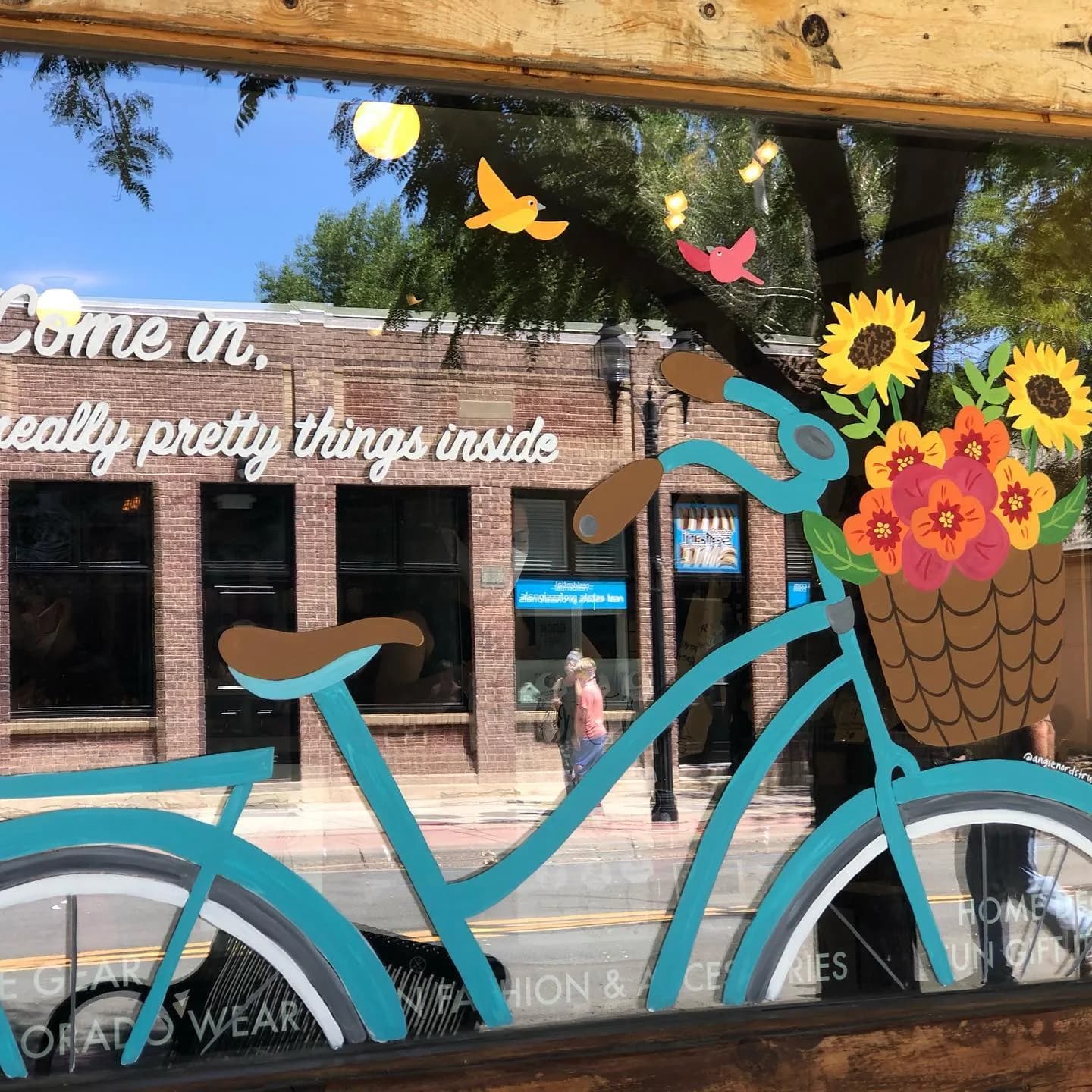 A large-scale painted mural depicts an oversized turquoise bicycle laden with flowers and flying birds against an urban brick storefront.