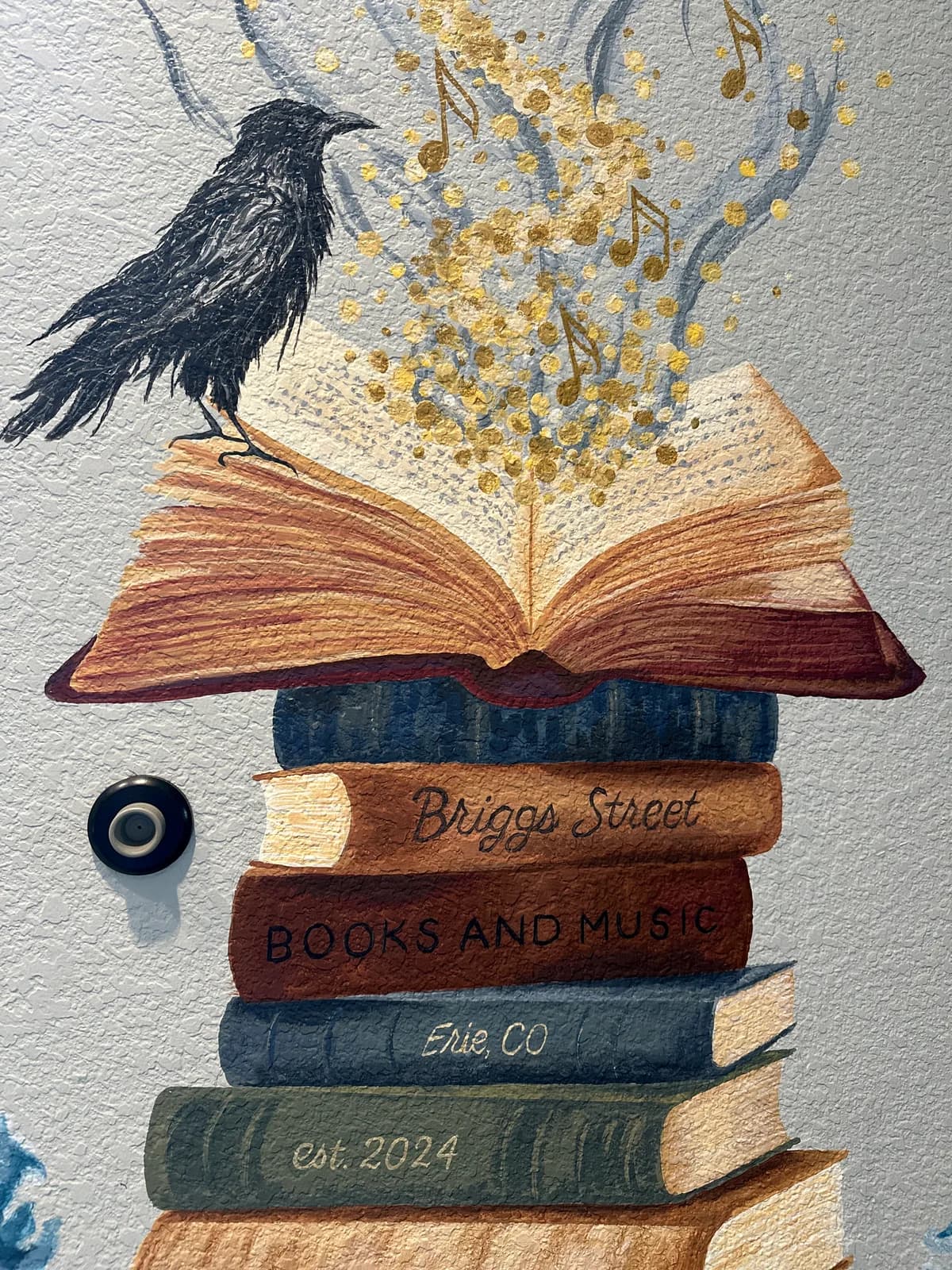 A dark raven perches atop stacked books with gold-leaf page details, literarily arranged above text identifying Briggs Street Books and Music in Erie, Colorado.