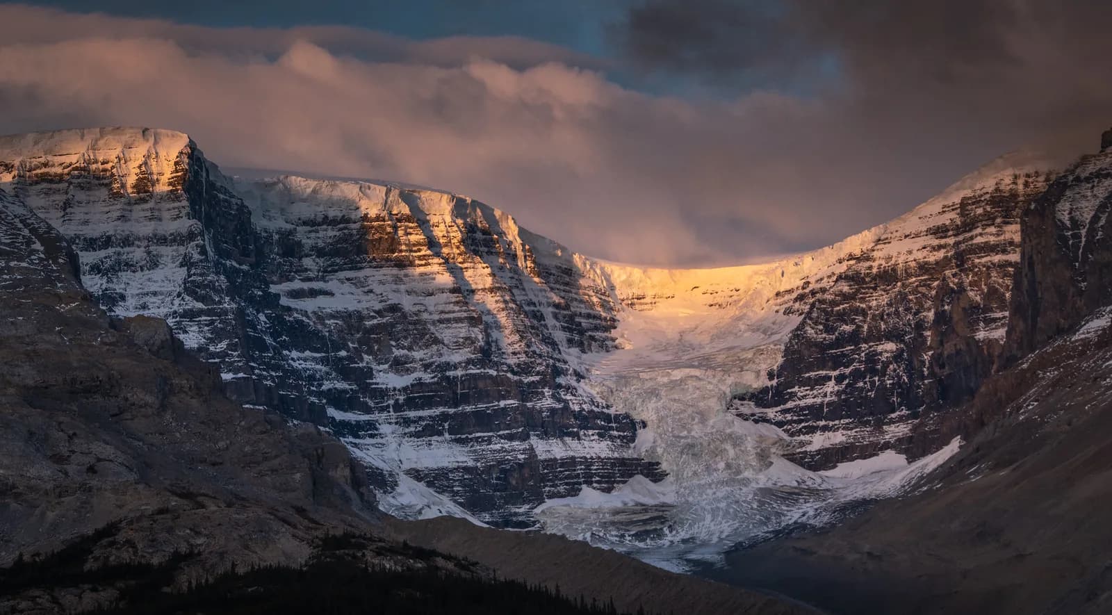 Snow-covered alpine peaks with a glacier-filled valley, illuminated by golden-hour sunlight against a moody sky.
