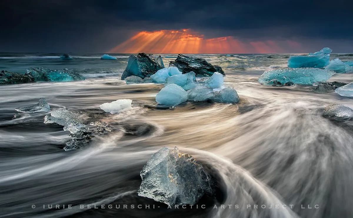 Glacial ice chunks and dark rocks on a beach under dramatic golden sunrays breaking through storm clouds.
