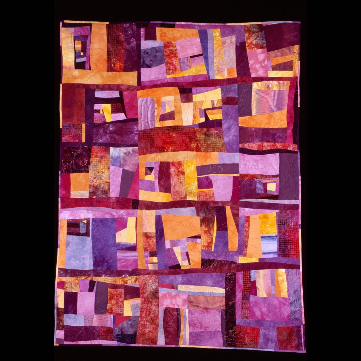 Abstract patchwork textile quilt composed of geometric fabric blocks in warm purples, oranges, and reds.