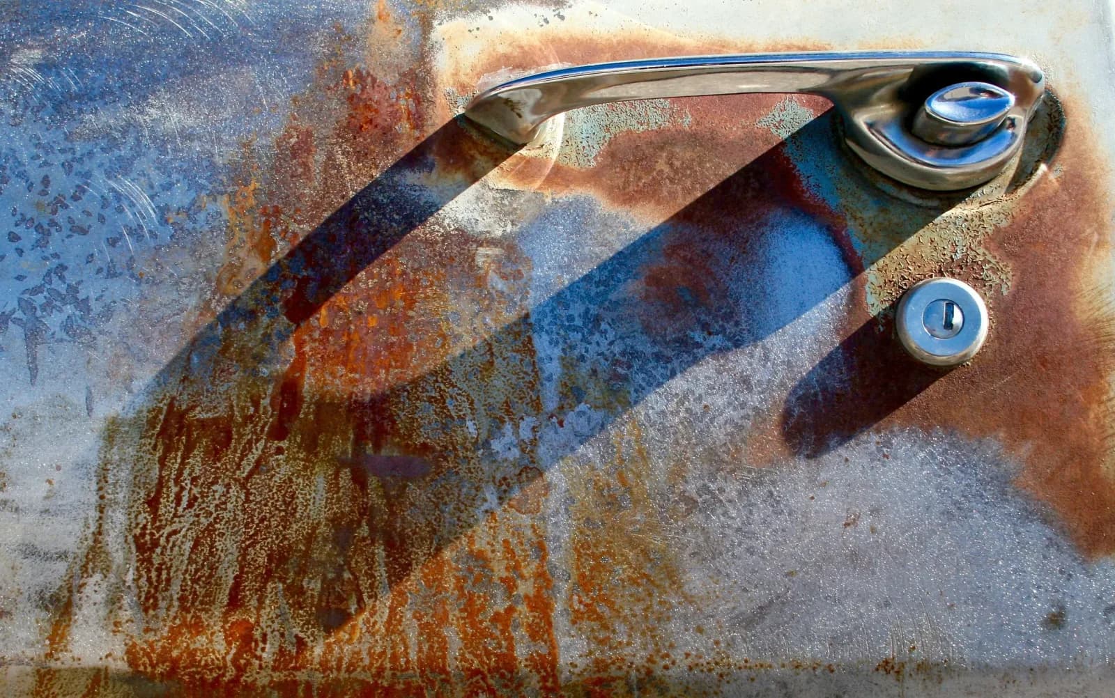 Rusted car door handle and trim detail against weathered, patina-rich metal surface, photographed in color.
