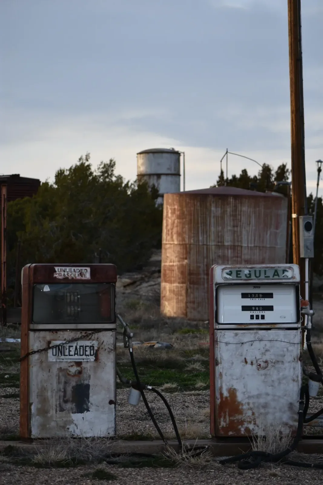 Two weathered vintage gas pumps stand abandoned in a high-desert landscape beneath a water tower.