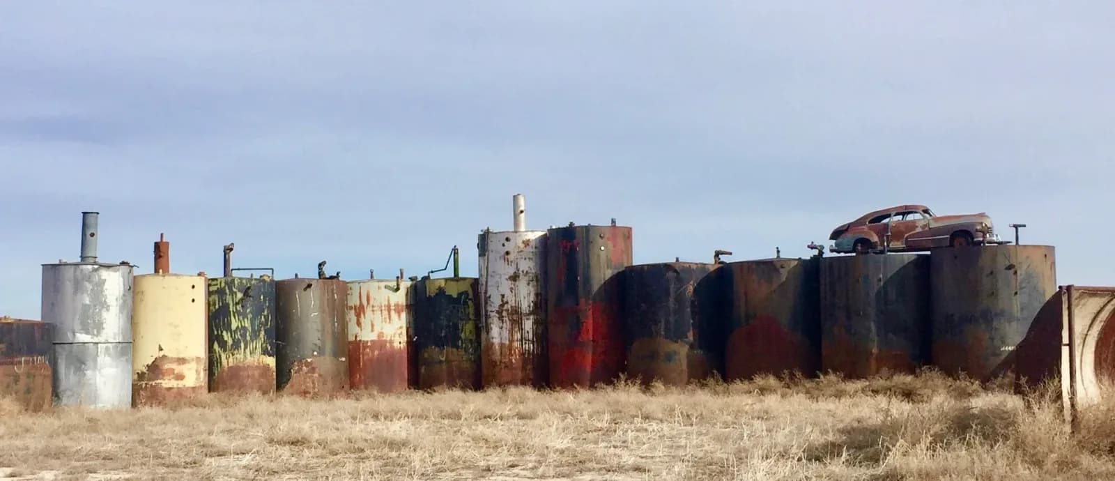 Row of weathered industrial tanks and abandoned structures in an open, arid landscape.