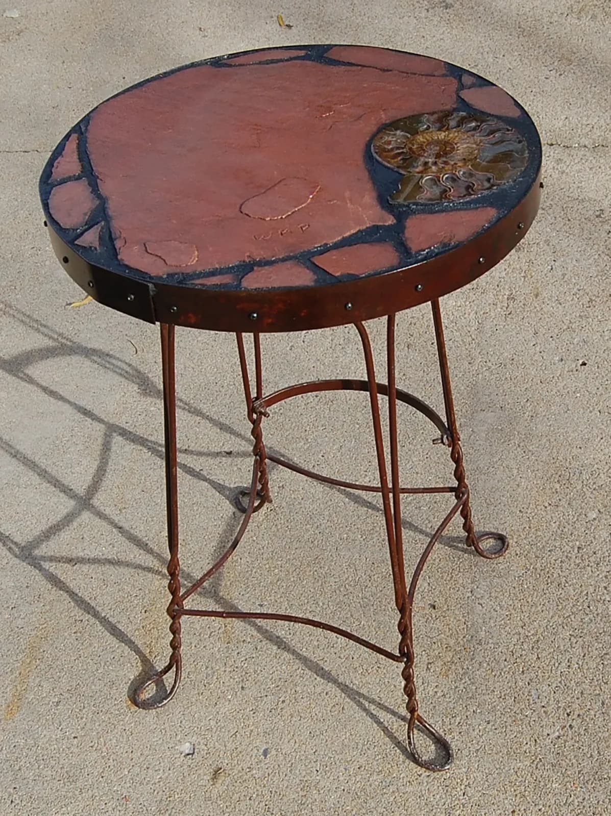 Mixed-media side table combining forged metal legs, red resin surface, and embedded fossil detail.