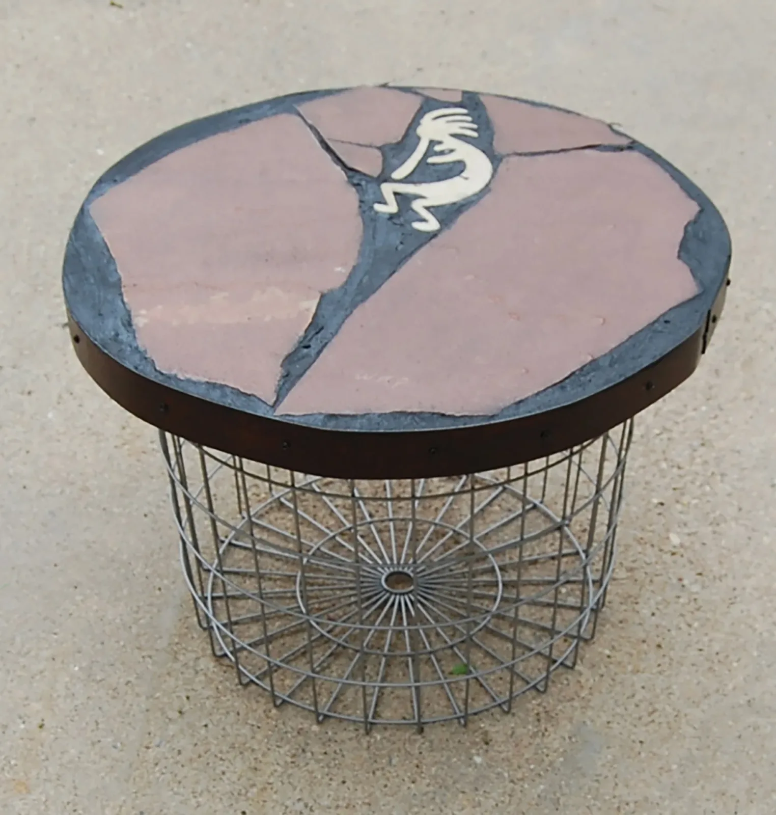 Hand-painted stone tabletop with kokopelli figure sits atop a wire basket base in mixed-media assemblage.