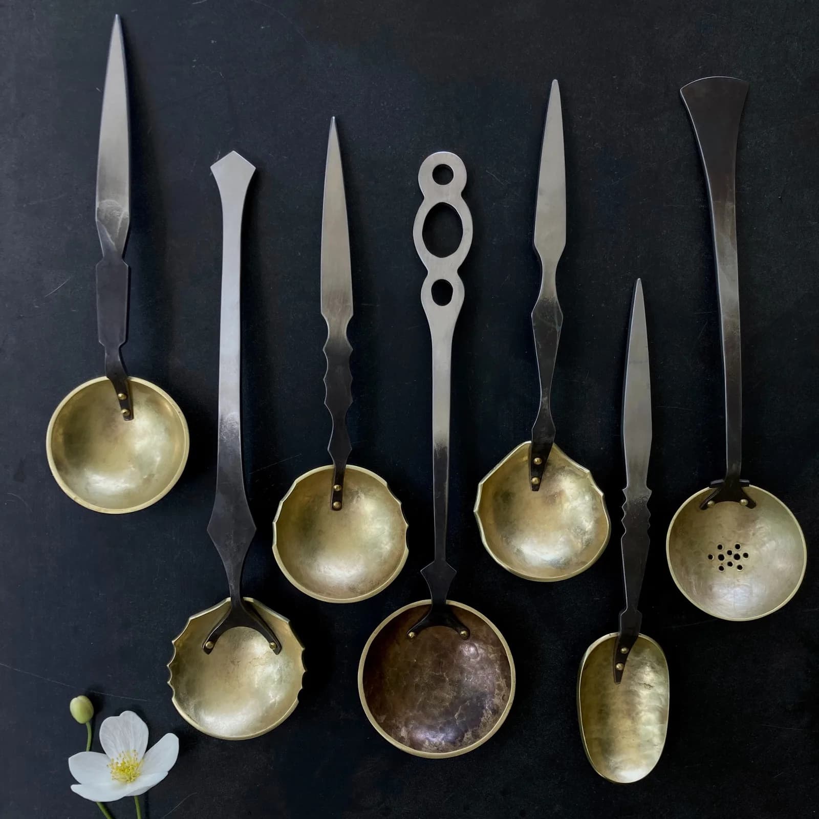 A collection of artisan brass and metal spoons with elongated handles and sculptural forms, arranged on dark backdrop.