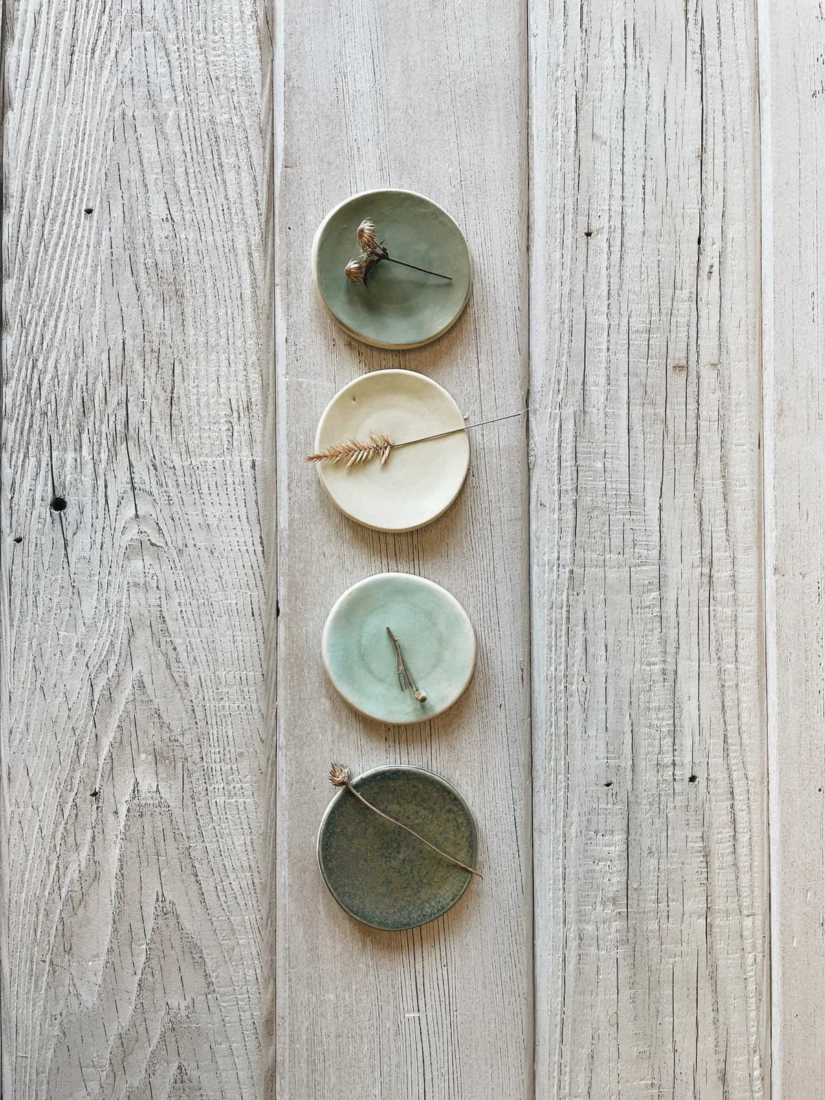 Four ceramic bowls in muted earth tones with delicate natural materials arranged vertically on weathered wood.