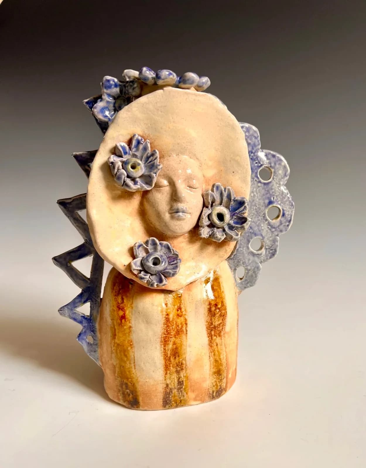 Ceramic sculpture of a stylized portrait head with blue flower and spiral elements.
