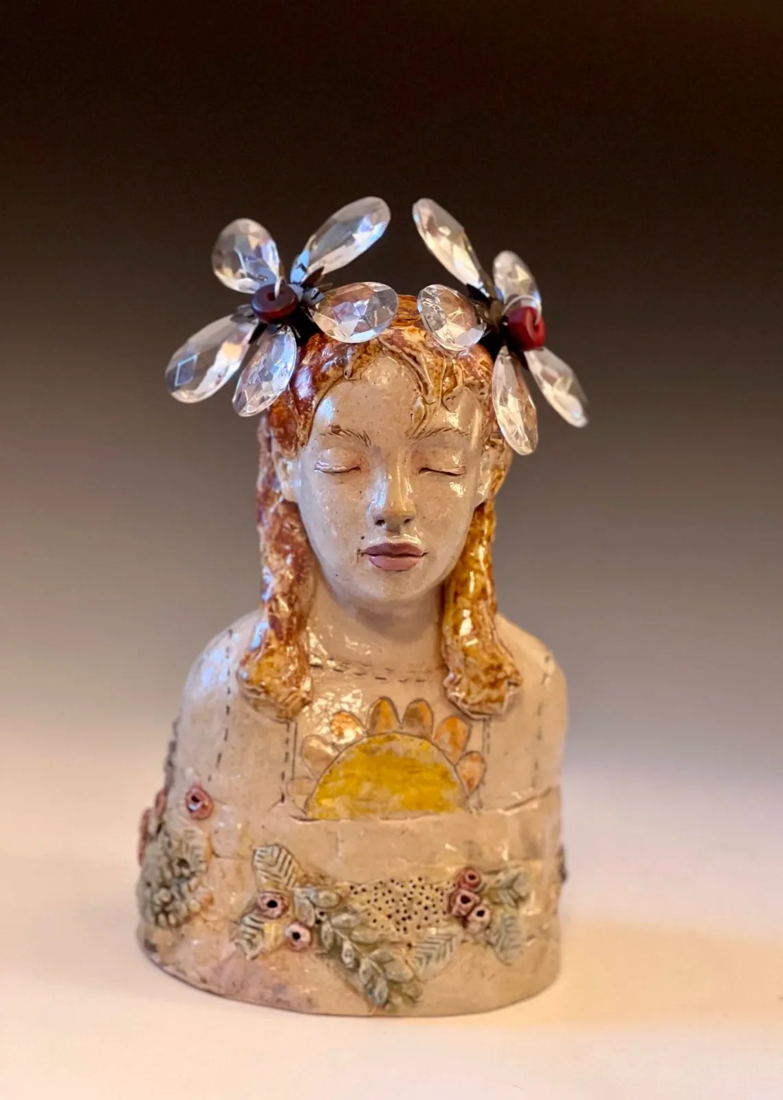 Ceramic portrait bust with blonde braids and glass flower crown, decorated with painted details and embedded jewels.