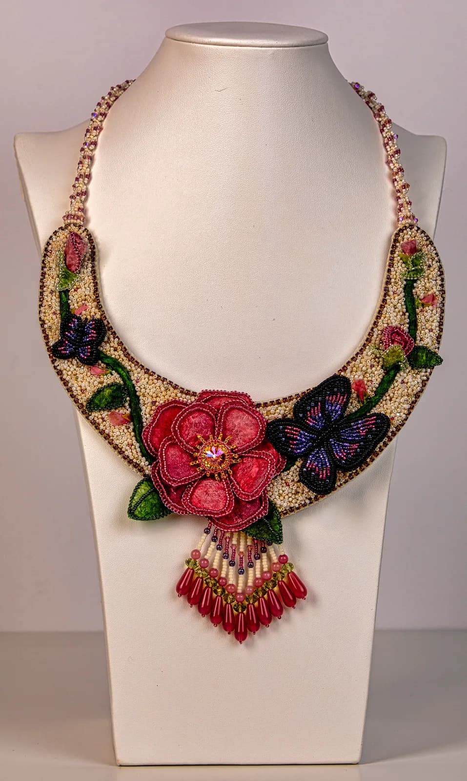 Beaded statement necklace with dimensional flowers, butterflies, and fringe in red, green, and gold tones.