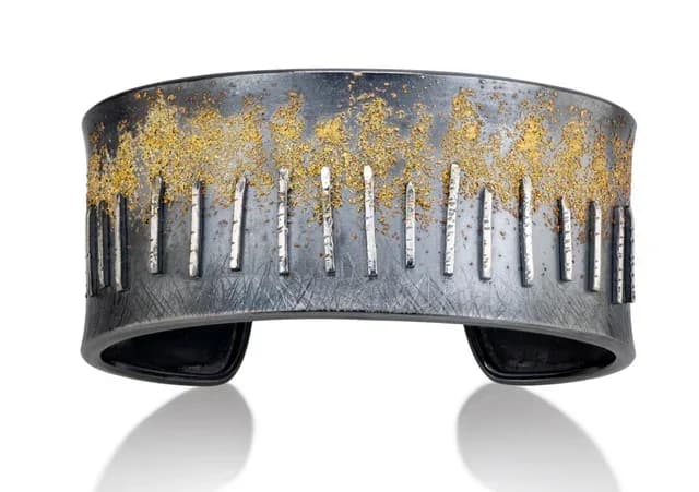 Oxidized metal cuff bracelet with gold-leaf accents and vertical slit details, photographed against white.