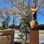 Abstract wooden sculpture mounted on a stone base, displayed outdoors in a garden setting.