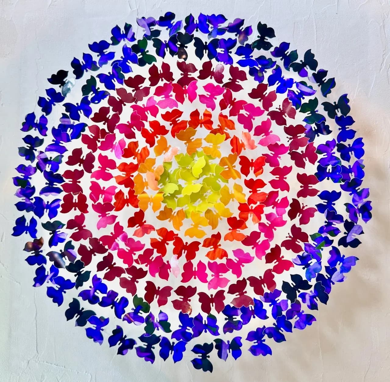 Circular composition of hundreds of colorful cut-paper butterflies arranged in concentric rings around a white wall.