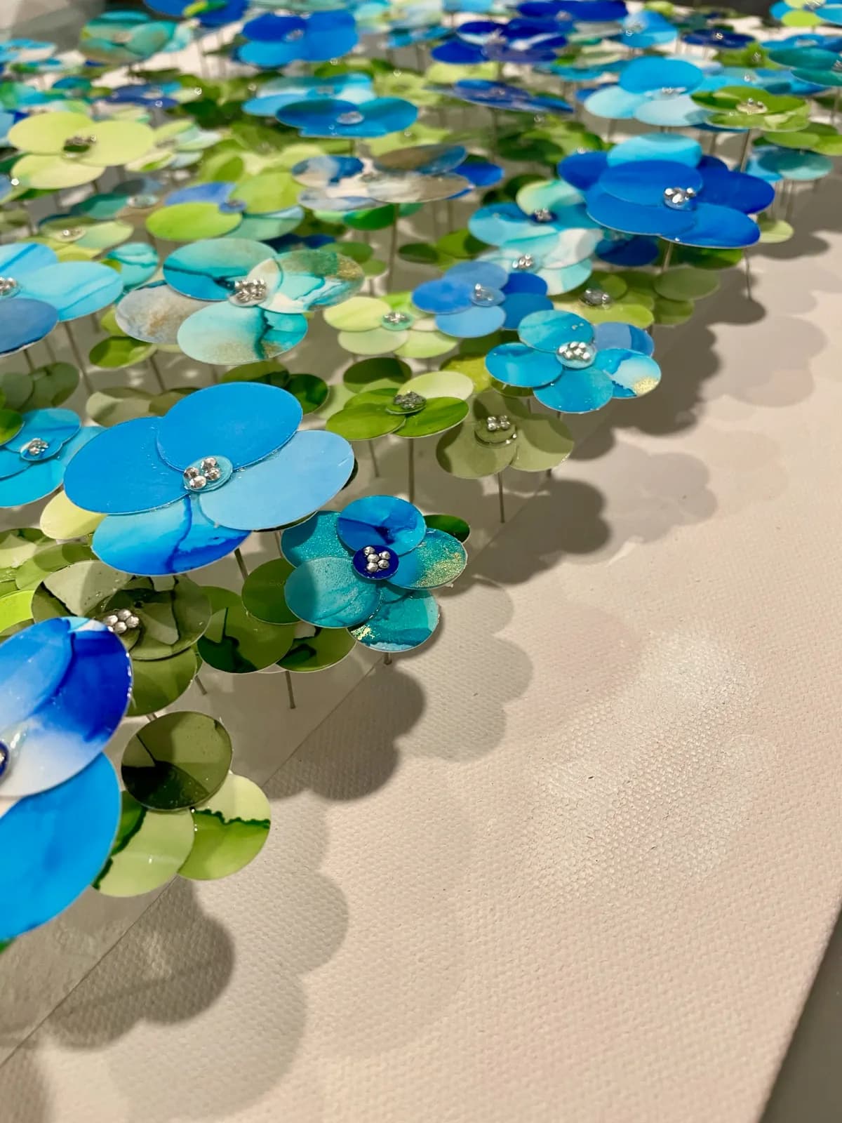 Three-dimensional metal flowers with blue, turquoise, and green petals adorned with jeweled centers cast shadows on white wall.