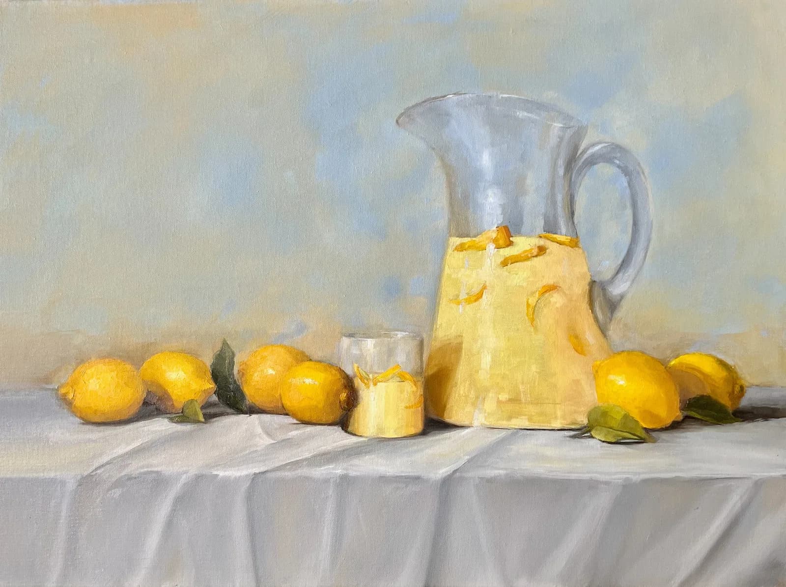 A clear glass pitcher filled with lemon water sits among scattered lemons and a drinking glass on a draped white tablecloth.
