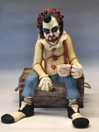 A ceramic figure of a clown-faced character in a cream and blue outfit, seated on a wooden log.