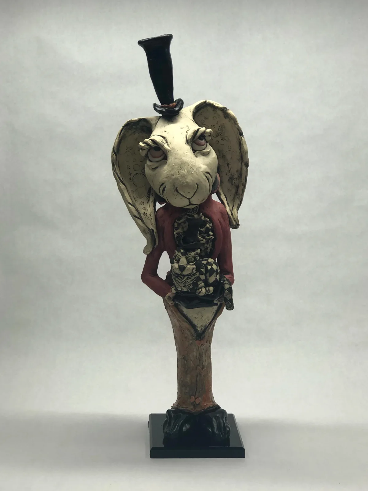 A surreal clay figurative sculpture combines a winged creature head, tailored jacket, and top hat on a pedestal.