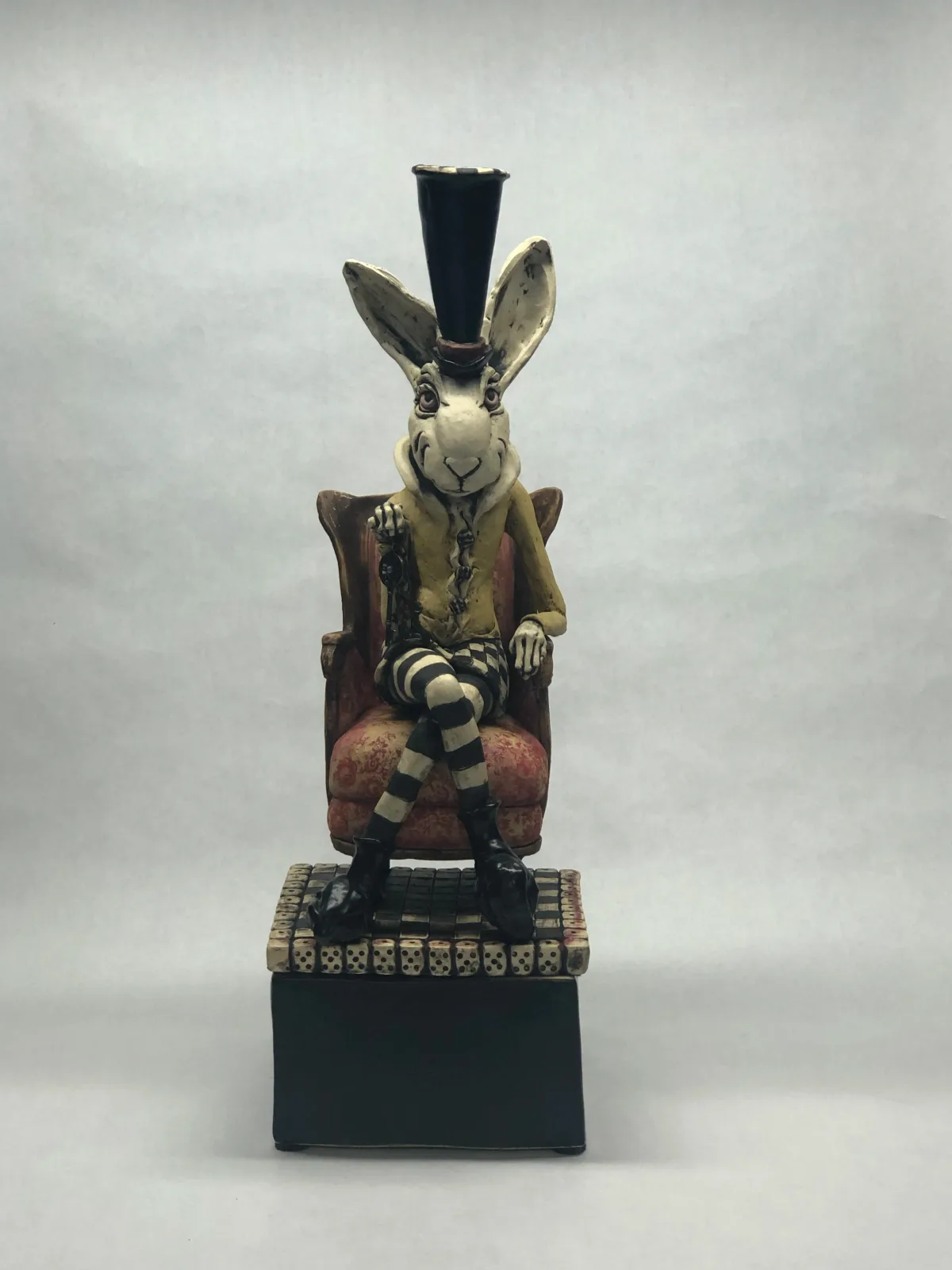 An anthropomorphic rabbit seated in a wooden chair wears a tall black hat and holds patterned fabric, crafted in wood.