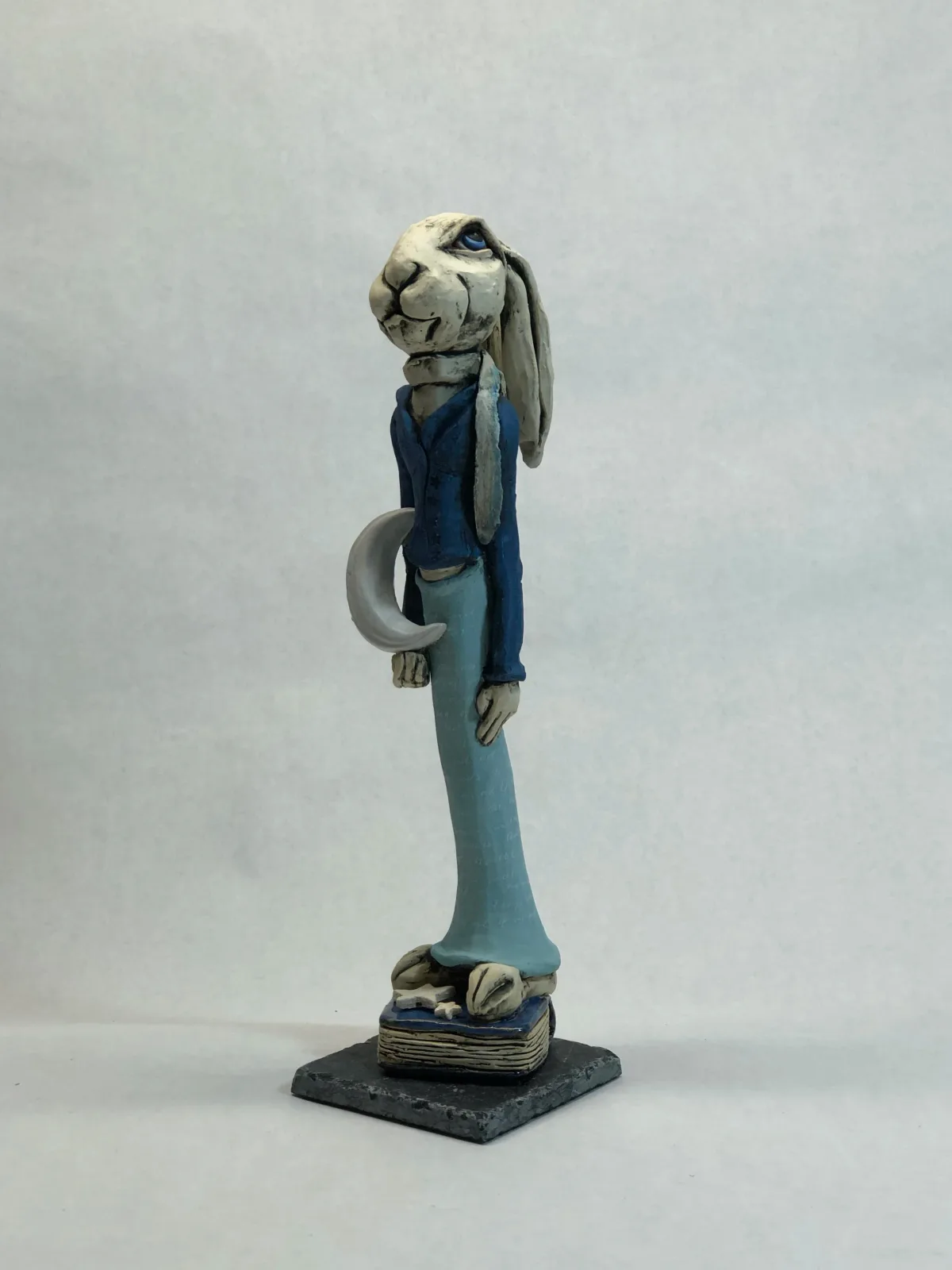 Ceramic figurative sculpture of an anthropomorphic goat in blue formal attire standing on a dark base.