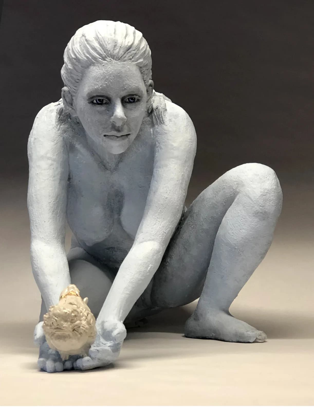 Seated figurative sculpture in gray clay with a small gold-toned object held in the subject's hand.