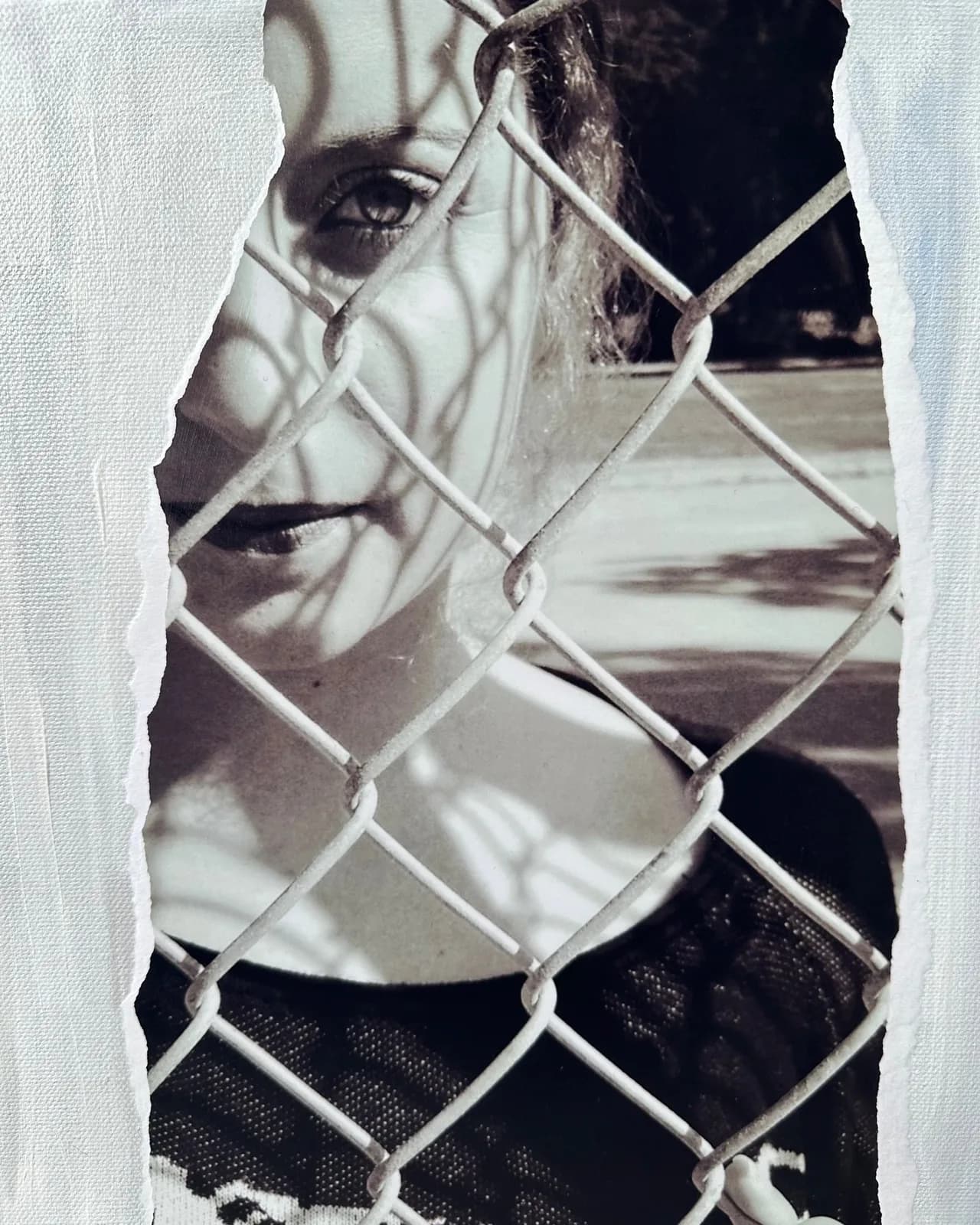 Monochrome photograph of a portrait viewed through chain-link fence, emphasizing confinement and shadow.