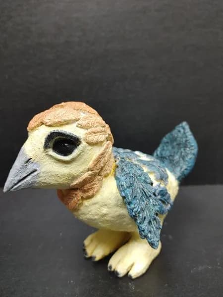 Small clay bird sculpture with tan head, black eye, and turquoise-blue feathered wings.