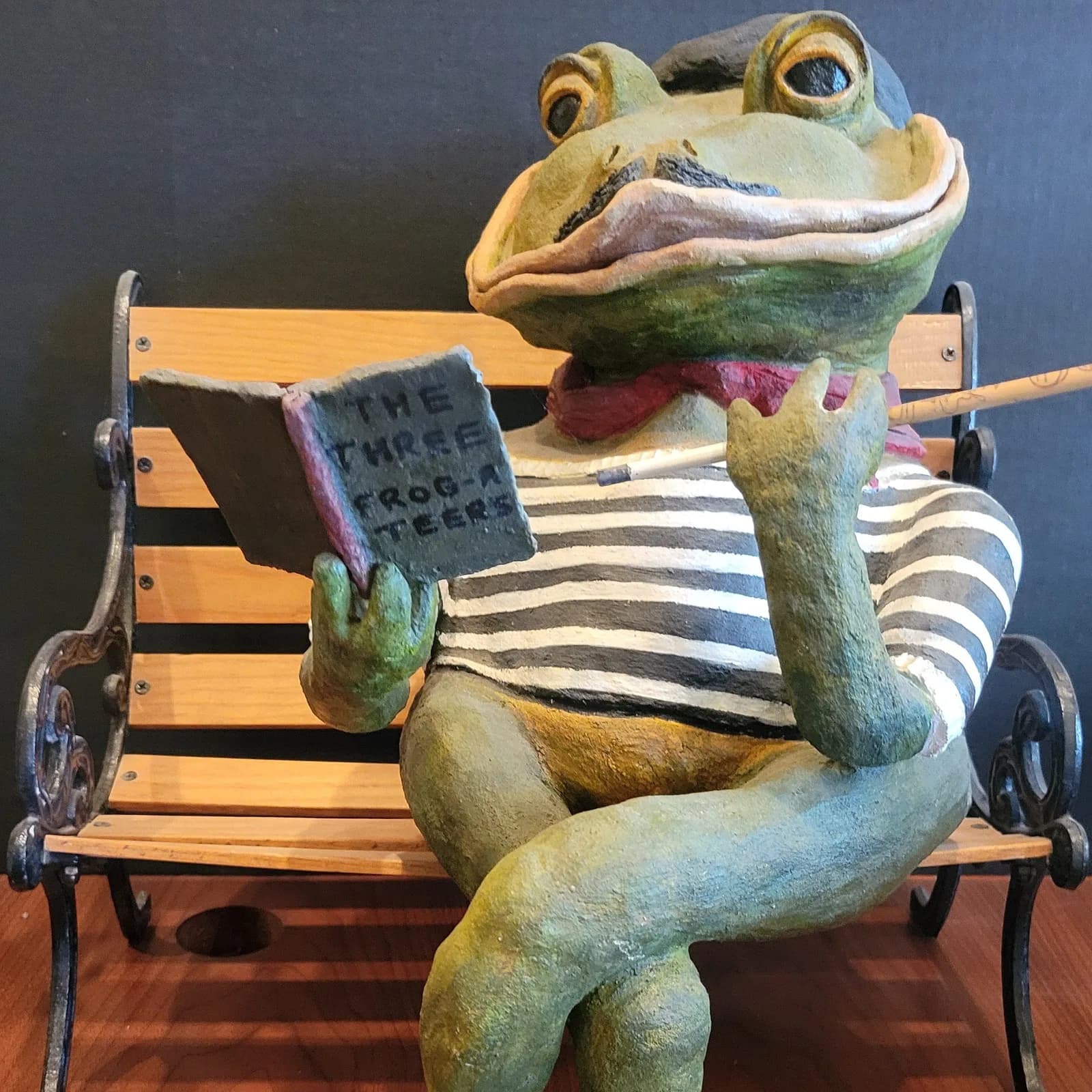 Whimsical carved frog reading a newspaper, seated on a striped wooden bench.
