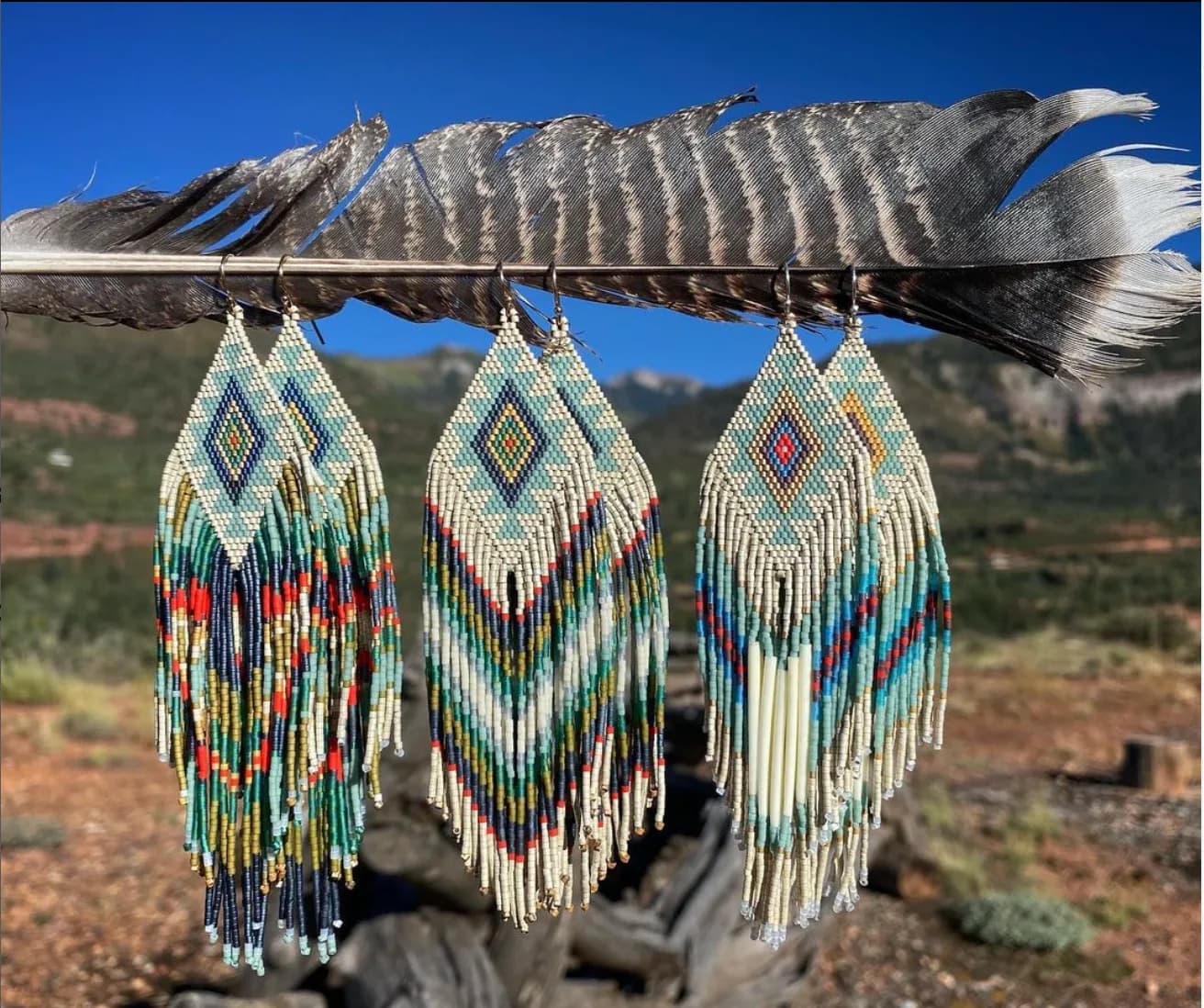 Three beaded feather ornaments with geometric patterns hang from an eagle feather above the Boulder Flatirons landscape.