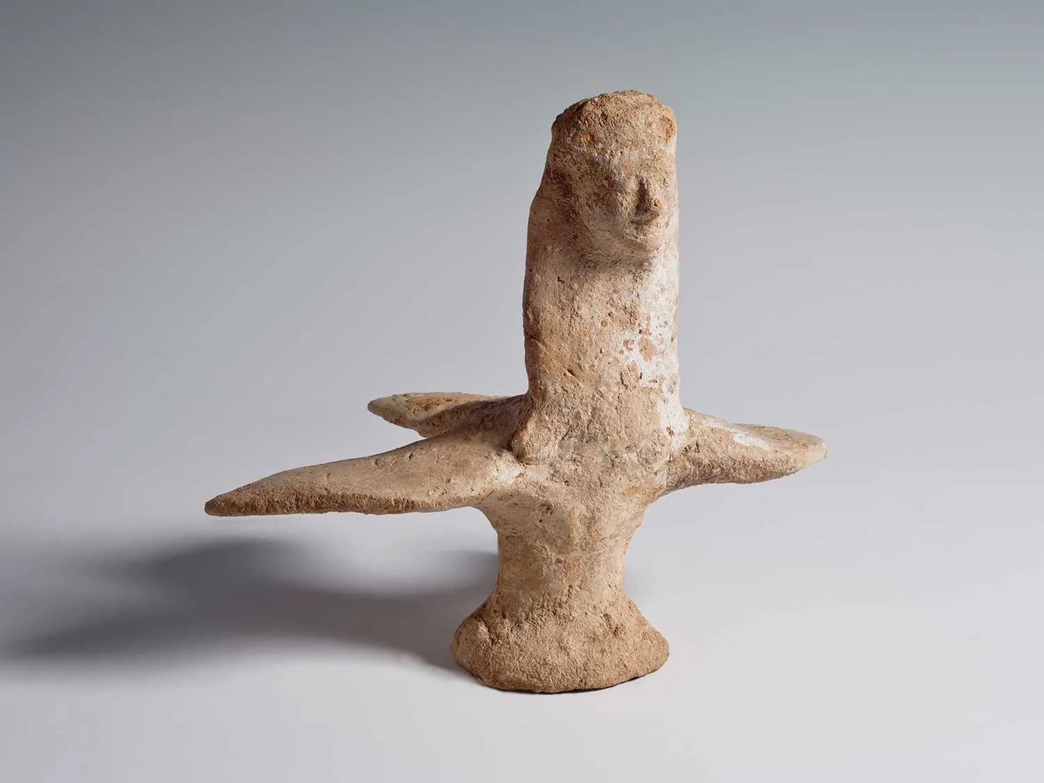 Abstract stone sculpture with elongated vertical form and horizontal arms, carved from warm tan stone.