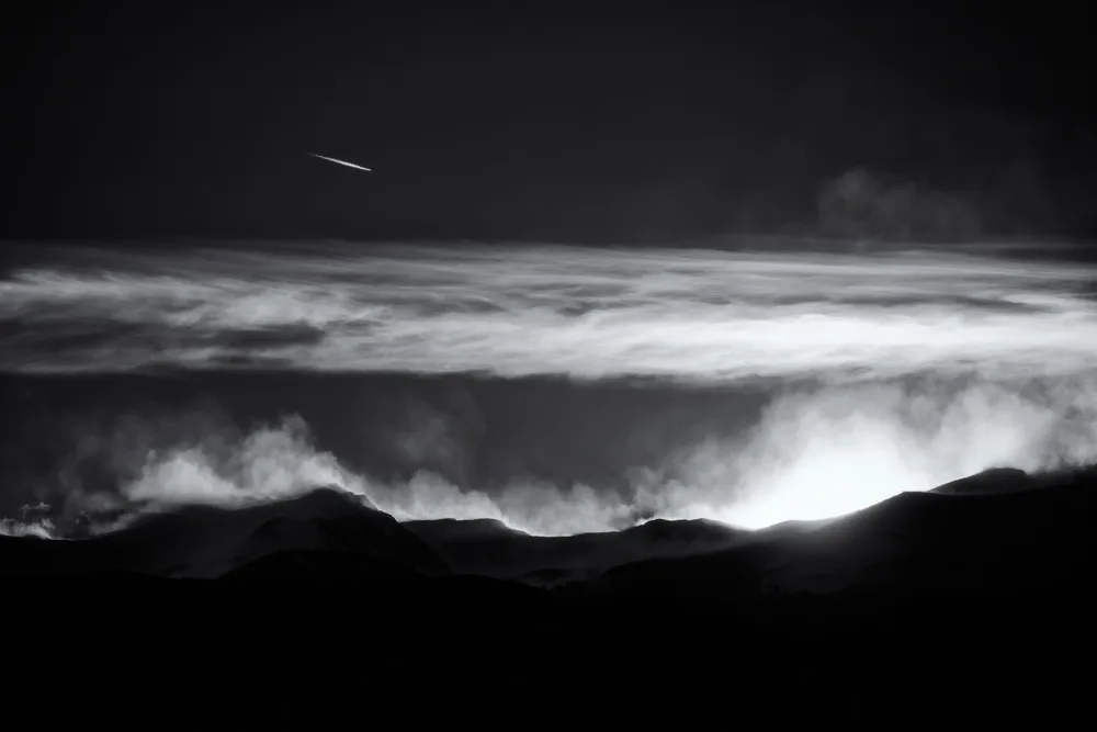 Black-and-white landscape photograph capturing mountains silhouetted against luminous cloud formations and dark sky.