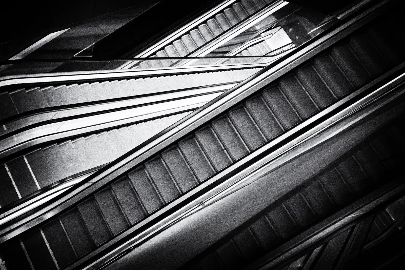 Monochrome photograph of escalator steps and handrails in geometric abstract composition.