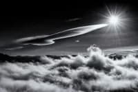 A dramatic black-and-white photograph capturing sunlight breaking through clouds from above, with an airplane silhouetted against the luminous sky.